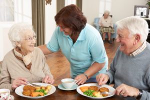 a carer helping an elderly man and woman at dinner a table. They are smiling.