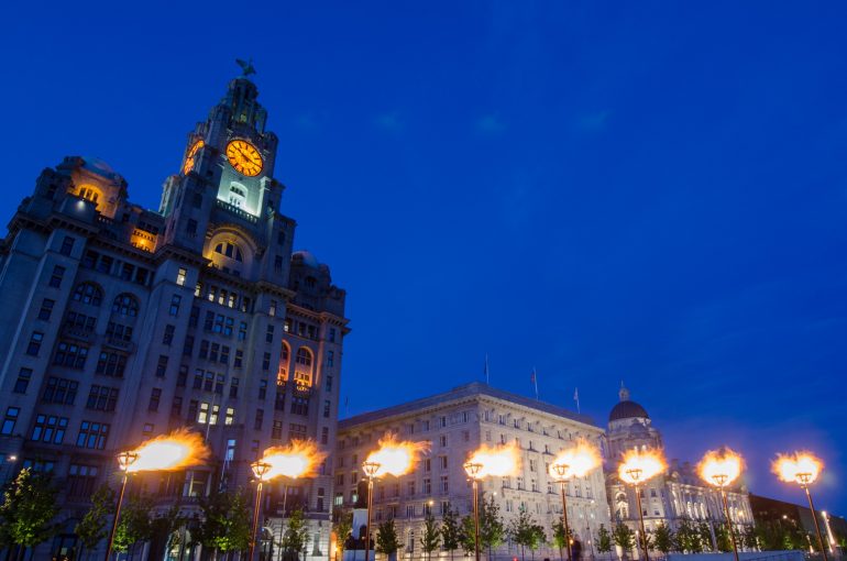 8 lit up beacons at night with the Liver Building and the Cunard Building in the background