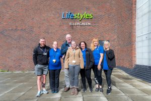 The image shows a group of seven people standing outside the Lifestyles Ellergreen centre, a brick building with the "Lifestyles Ellergreen" logo displayed on the wall behind them. The group consists of a mix of men and women, some wearing Lifestyles uniforms in blue and black, and a few in casual clothing. They are smiling and standing close together, with one of the men, an older individual, having his arm around a younger woman in the centre. The mood is positive, and the group appears united, likely representing the Lifestyles staff and a member.