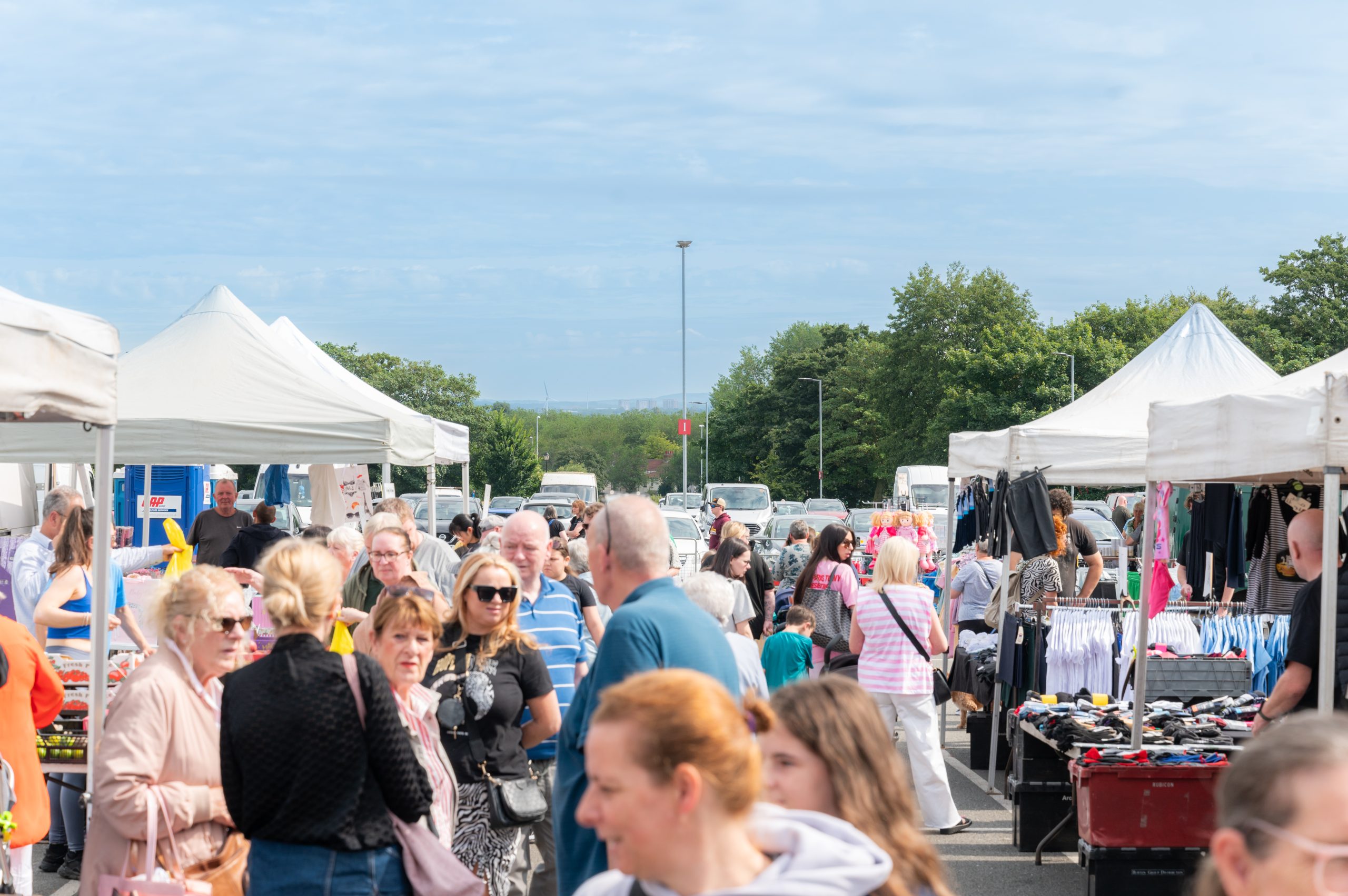 This image depicts a bustling outdoor market on a sunny day. A diverse crowd of people is walking between rows of white-tented stalls that line both sides of the scene. Some people are engaged in conversation, while others are browsing the various goods on display, including clothing and other items. The market appears to be well-attended, with a mixture of ages among the attendees. In the background, there are parked cars, trees, and a clear, blue sky with a few scattered clouds, giving the setting a lively yet relaxed atmosphere.