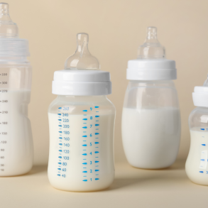 "Four baby bottles filled with milk, varying in size and measurement markings, displayed on a neutral beige background."