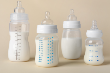 "Four baby bottles filled with milk, varying in size and measurement markings, displayed on a neutral beige background."