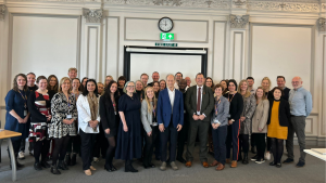A group of Liverpool City Council staff members posing for a photo with Professor Sir Michael Marmot, who stands in the center wearing a blue suit jacket and white shirt. The group is gathered in a bright, formal room with ornate detailing on the walls, a clock, and an exit sign above a screen in the background