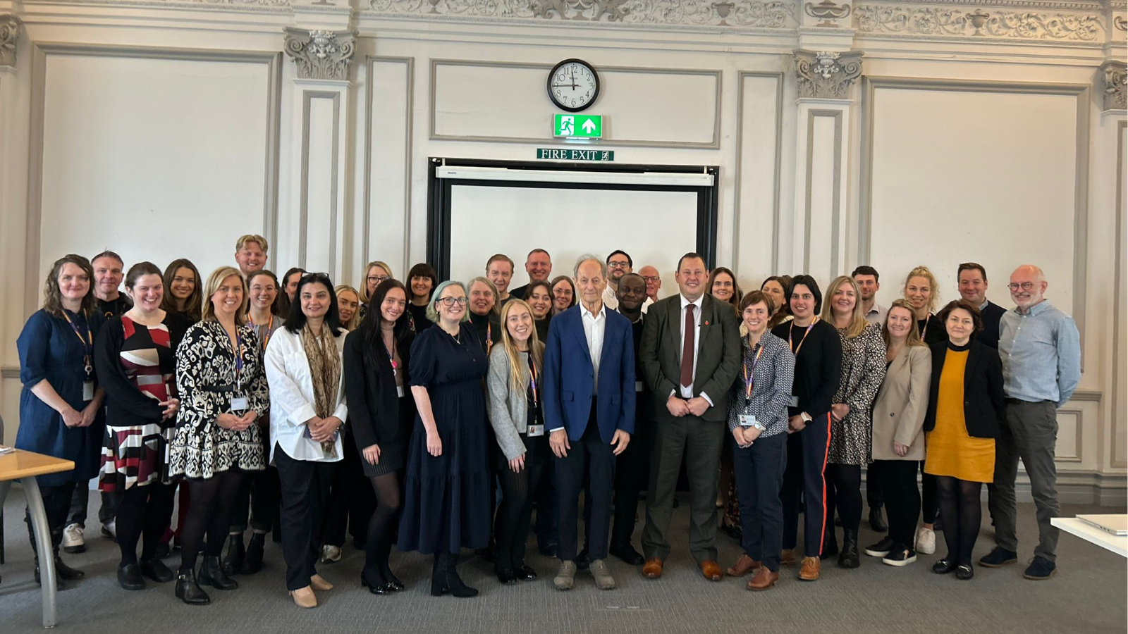 A group of Liverpool City Council staff members posing for a photo with Professor Sir Michael Marmot, who stands in the center wearing a blue suit jacket and white shirt. The group is gathered in a bright, formal room with ornate detailing on the walls, a clock, and an exit sign above a screen in the background