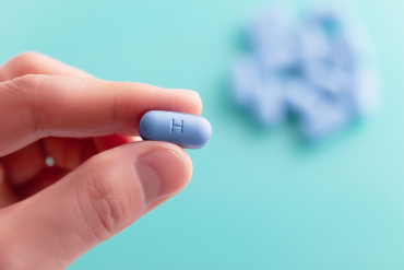 A close-up image of a hand holding a single blue pill with the letter "H" engraved on it, set against a soft teal background. In the blurred background, additional blue pills are visible.