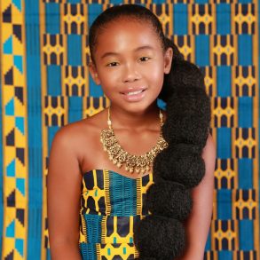 A young Black girl with long hair wearing a blue, yellow and black dress against a blue, yellow and black backdrop