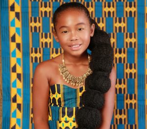 A young Black girl with long hair wearing a blue, yellow and black dress against a blue, yellow and black backdrop