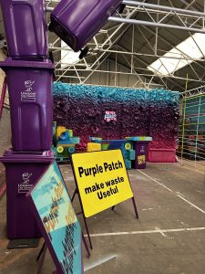 A metal sign for Purple Patch, with a wall of recycled rubbish spray painted purple behind it.