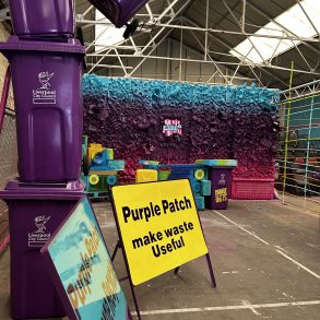 A metal sign for Purple Patch, with a wall of recycled rubbish spray painted purple behind it.