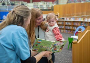 A toddler being shown a book by an adult whilst her mother looks on